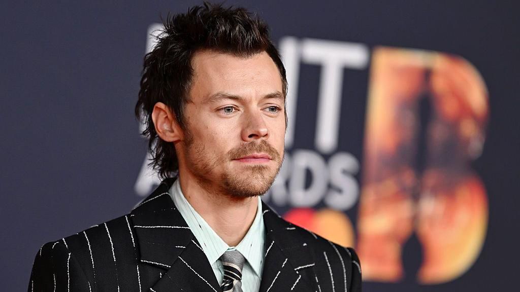 Harry Styles wears a pinstripe suit with a light green shirt and silver tie underneath. He stands against a Brits backdrop
