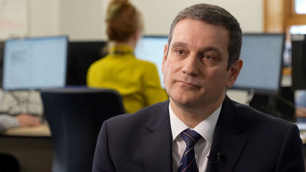 Stephen Clayman, with short brown hair, wears a dark blue suit with a white shirt and striped navy tie. In the background blurred police officers in plain clothes can be seen sat at work stations.