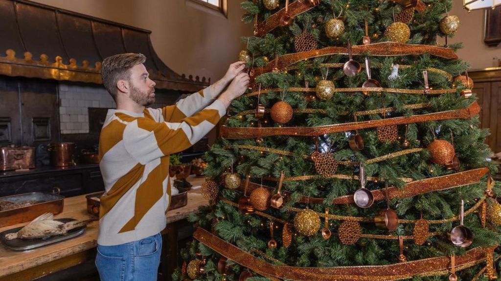 Man decorating a tree in a kitchen with copper ribbons and coper pots and ball-balls 