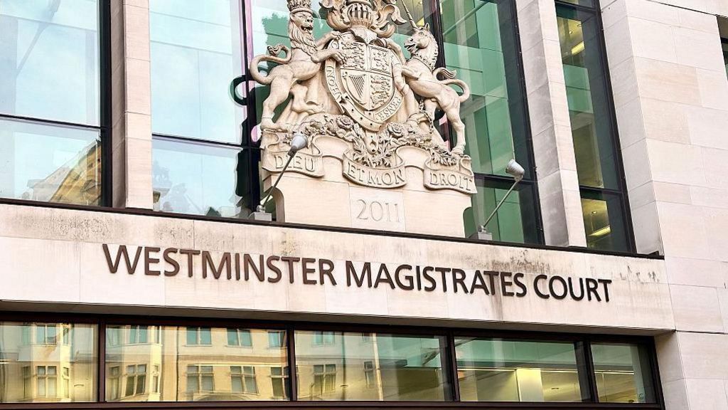 The outside of Westminster Magistrates Court. It has glass windows which have a reflection of buildings in them. And the sign above the windows says 'Westminster Magistrates Court', which is below a historical crest.