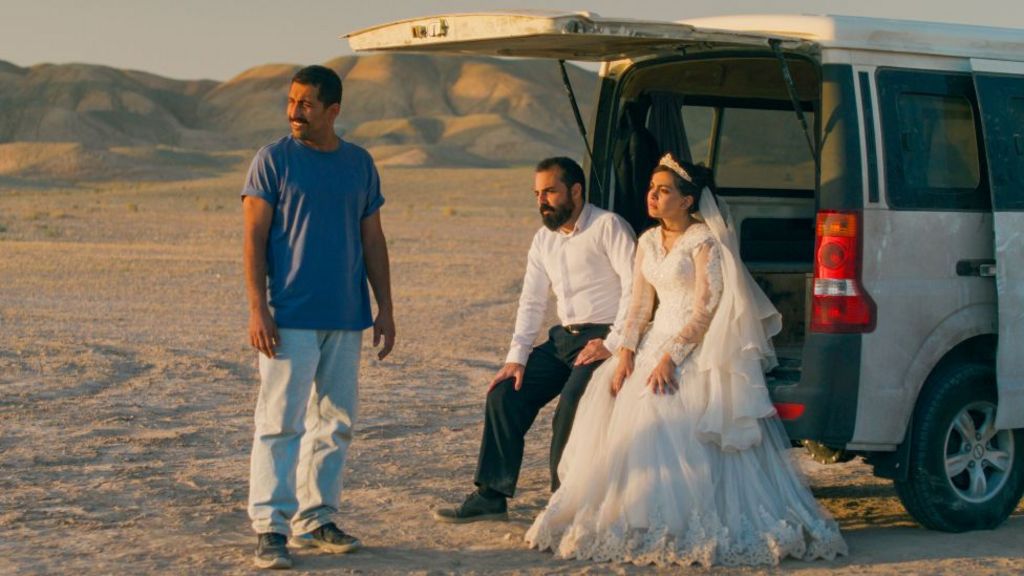 A bride and groom sitting in the back of a car with its boot open, while another man stands beside them, in a promotional image from It Was Just An Accident