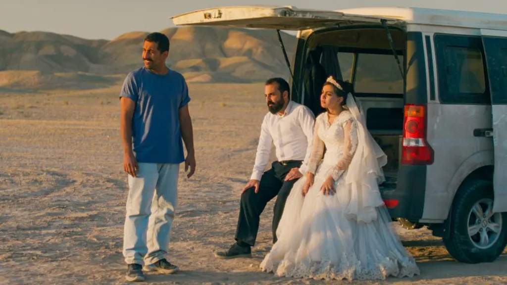 A bride and groom sitting in the back of a car with its boot open, while another man stands beside them, in a promotional image from It Was Just An Accident