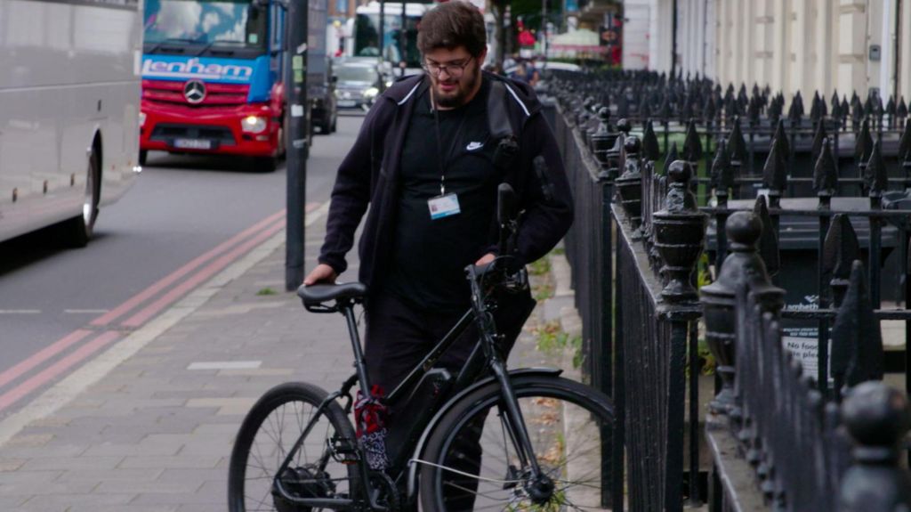 Bilal, dressed in black, holds a bike next to a road and black spikey railings