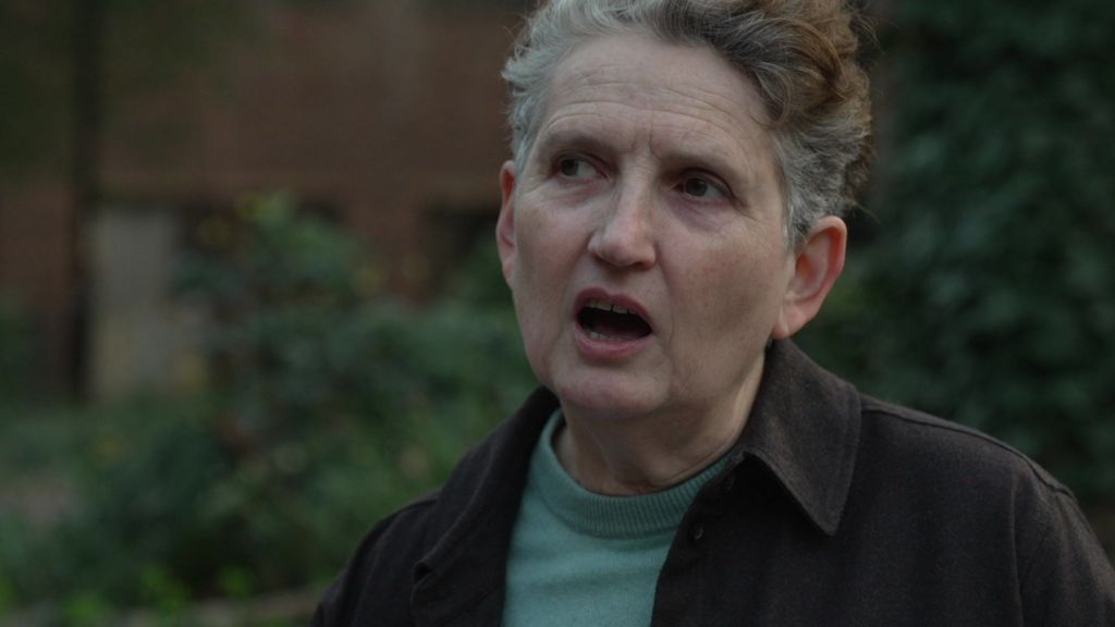 A middle-aged woman with short, greying curly hair and a brown shirt