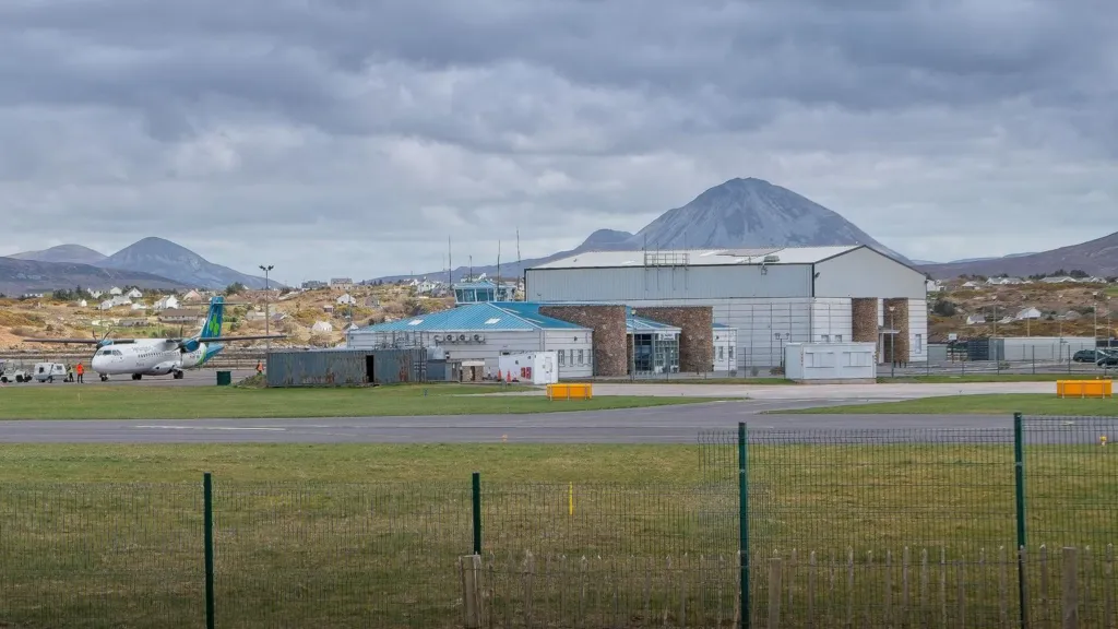 Carrickfinn Airport in County Donegal. An Aer Lingus plane sits on the runway near the airport, a small terminal building with a blue roof is to the right and beside that is a large hangar. Mountains can be seen in the background.