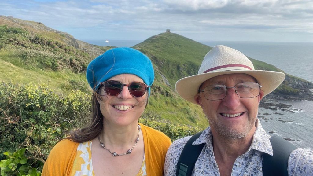 A smiling selfie of Lizzy Stroud and husband Dave standing high on a cliff with the sea and hills behind them. She is wearing a yellow cardigan and blue beret and sunglasses. He is wearing a pale blue floral shirt a sunhat, glasses and backpack
