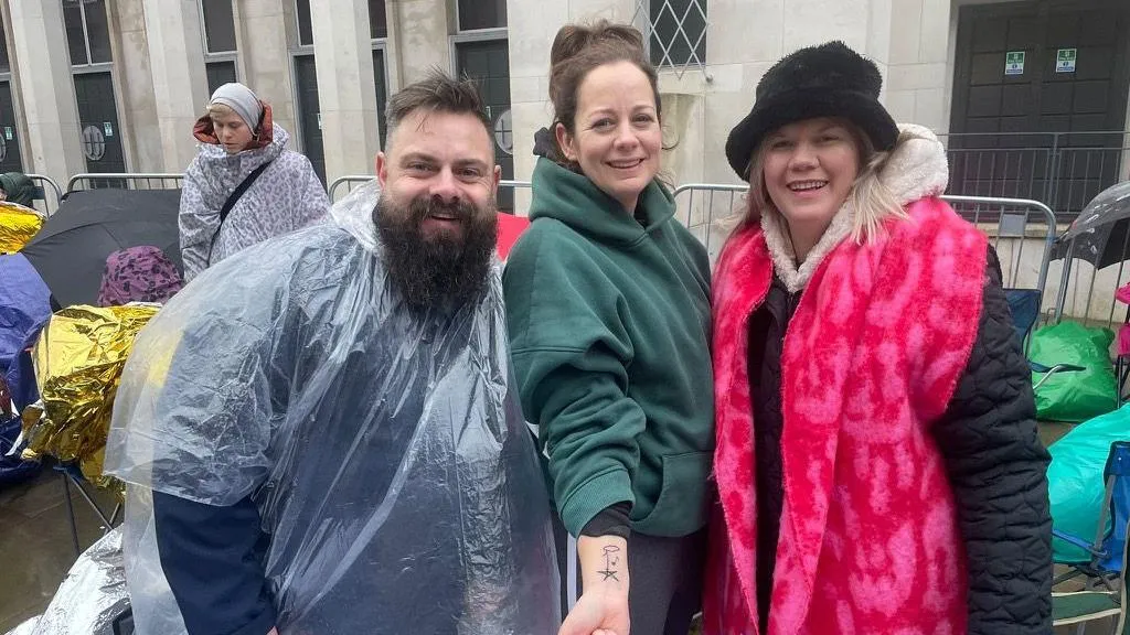 Scott White, Carla Hill, Danielle Elkington - a man in a see-through poncho is standing with two women, one is wearing a green hoody and the other is wearing a black hat, pink gilet over a black coat