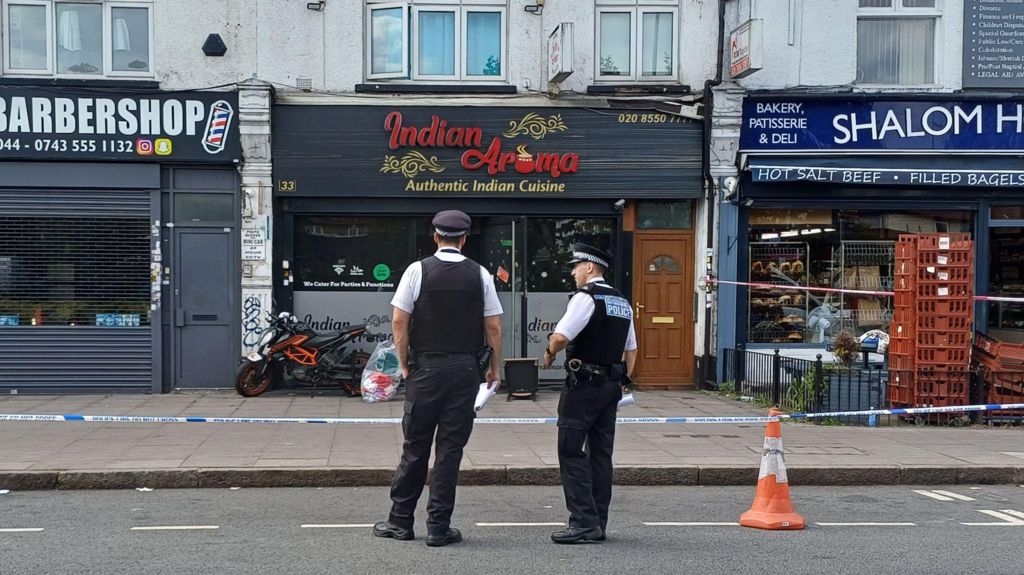Two police officers in uniform stand on a road in front of police tape which surrounds shops and restaurants. Their backs are to the camera. Signage for the businesses pictured indicate they are a barbershop, an Indian restaurant and a deli.