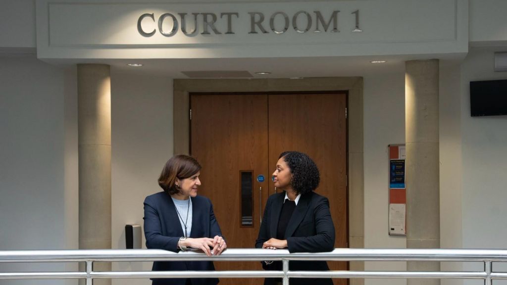 Two women in black suit jackets talk on a balcony with courtroom signage above their heads. Brown double doors can be seen in the background. 