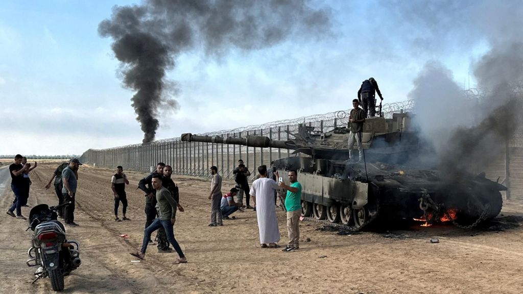 Palestinians climb on top of an Israeli military tank set damaged during Hamas's 7 October 2023 attack on Israel, on the Israeli side of the Israel-Gaza perimeter fence