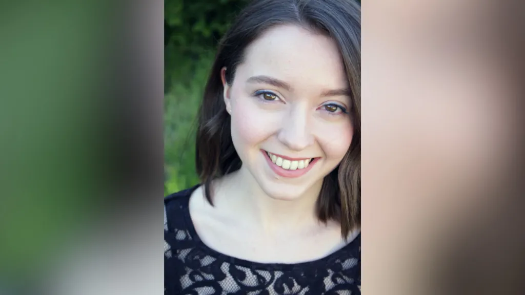 Lisa is outside. She has a brown bob and is wearing a black top. Some foliage is behind her. 