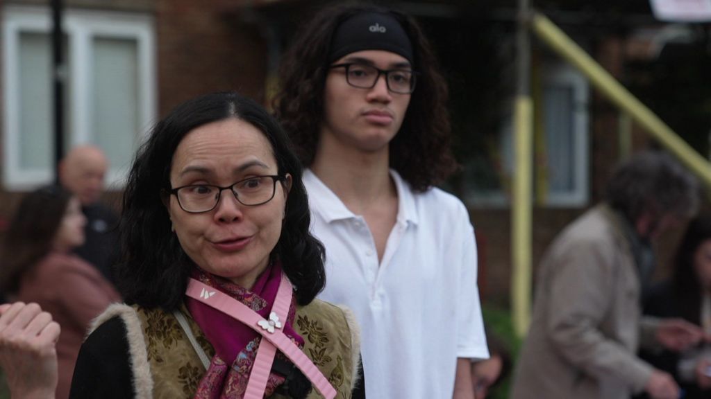 A middle-aged Chinese woman wearing glasses talks to camera with a teenaged boy with long hair next to her