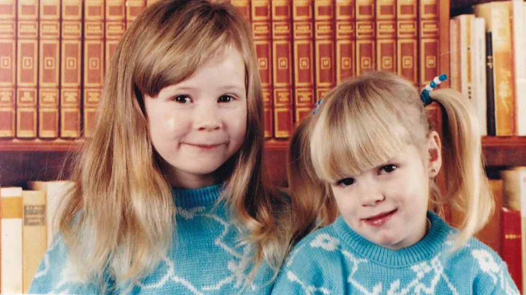 Hayley and Kirsty Wilkinson are two white girls aged approximately five and three. Hayley, left, has chest length blonde hair with a long fringe brushed to the side. Kirsty has blonde hair worn in two bunches. They are wearing identical turquoise jumpers with irregular white lines forming patterns on it. They are both smiling at the camera standing in front of bookshelves.