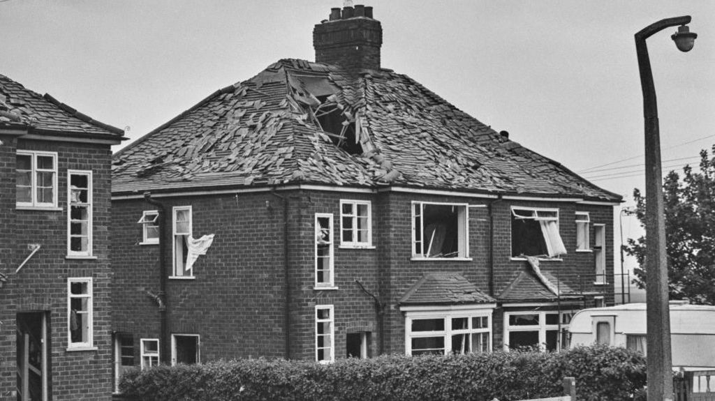 Black and white image of semi-detached houses with tiles stripped of roofs and a hole in one of them