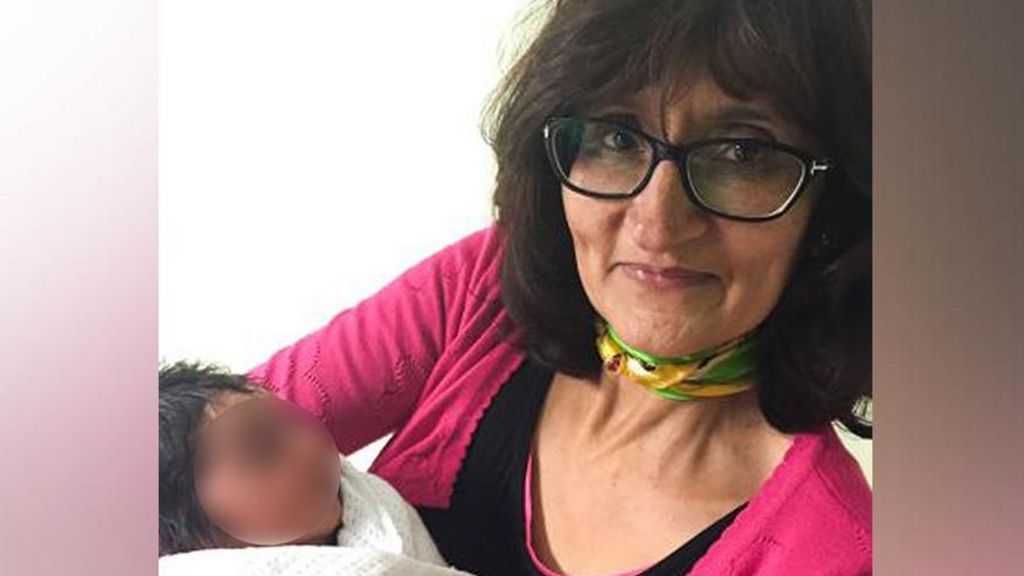 A woman with dark hair, glasses, wearing a pink cardigan holds a small baby in her arms. 