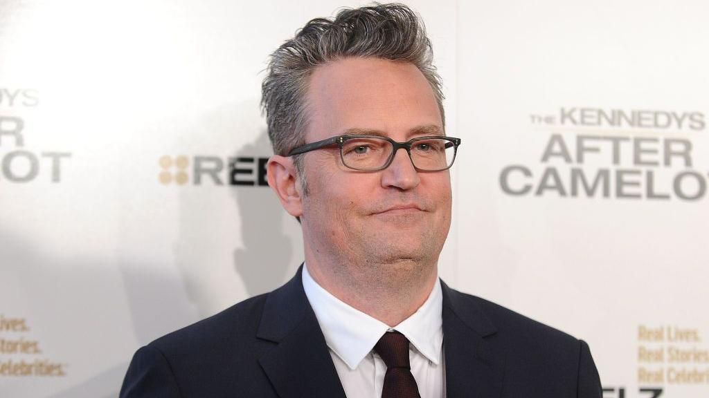 Matthew Perry wears glasses and a dark suit. He is posing for a picture on an event red carpet.
