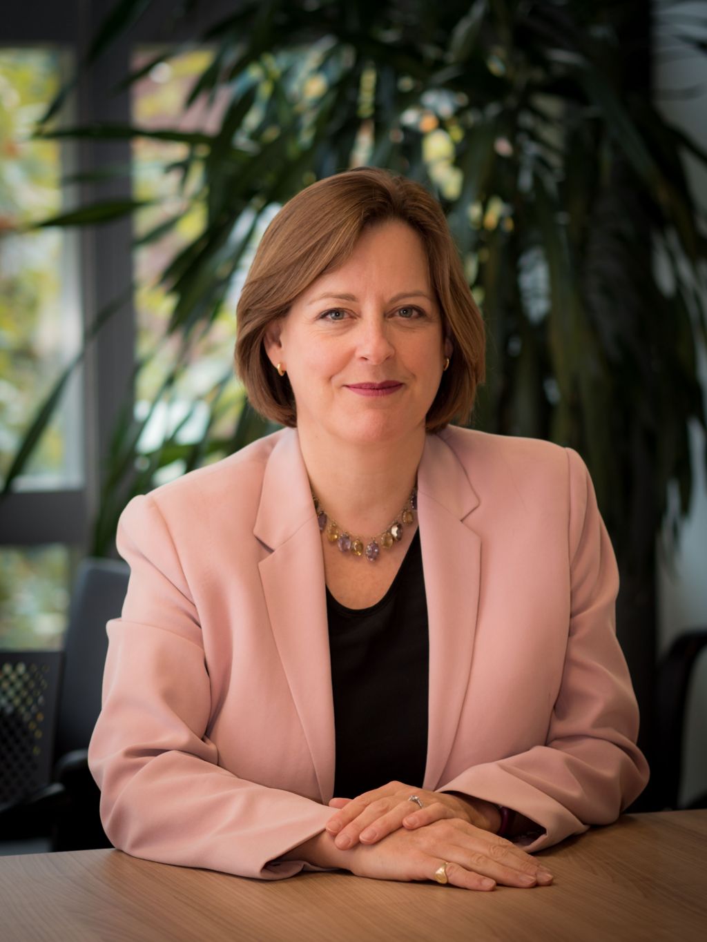 A woman in a salmon pink blazer sits at a table in a professional looking headshot style photograph