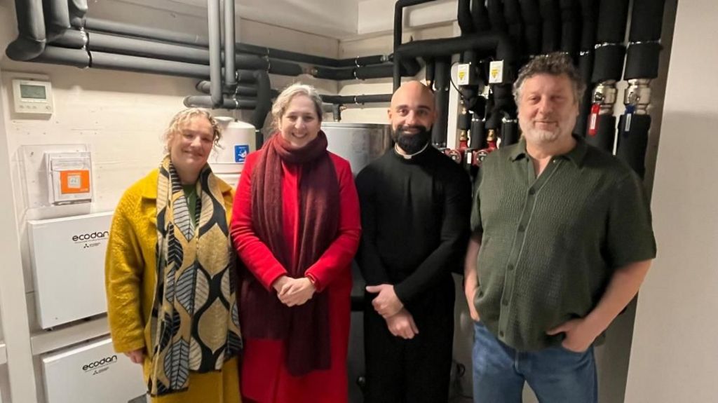 Four people stand in a row with pipes in the background. The third person along is a priest wearing his uniform with a dark beard and bald head. All smile at the camera
