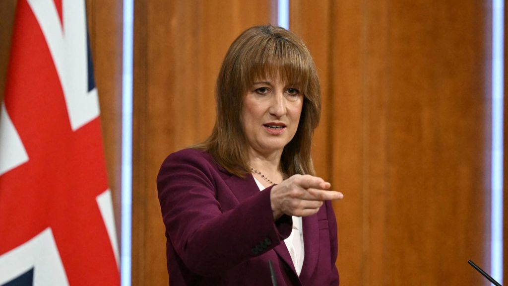 Rachel Reeves points as he takes questions from reporters, with a union jack flag in the background