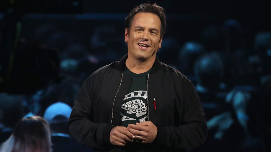 A man wearing a bomber jacket over a black t-shirt with white print stands on stage addressing a crowd