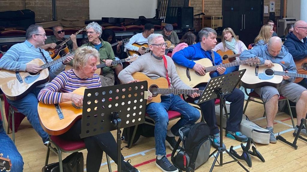 An ensemble of guitarists of all ages sit in two lines at a club with music stands in front of some of them.