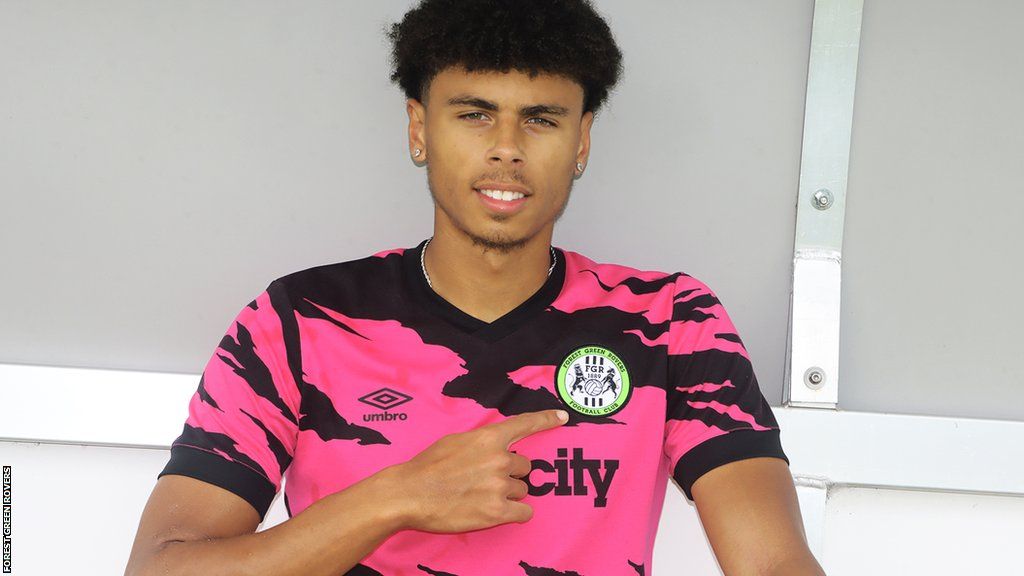 Reece Welch points to the Forest Green Rovers badge while wearing a pink and black shirt after signing for the League Two club