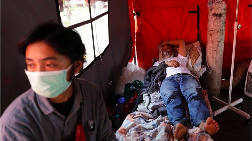 Ketut Nomer, a 59-year-old patient suffering from coronavirus disease (COVID-19), rests as his 28-year-old son Gede Zico sits taking care of him, at a temporary tent outside the emergency ward of a government hospital in Bekasi, on the outskirts of Jakarta, Indonesia, 25 June 2021.