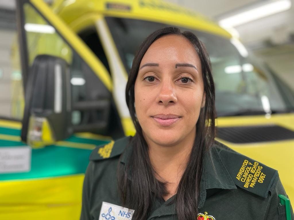 Tamara is wearing the green ambulance service uniform and is stood in front of an ambulance. She has long dark hair.
