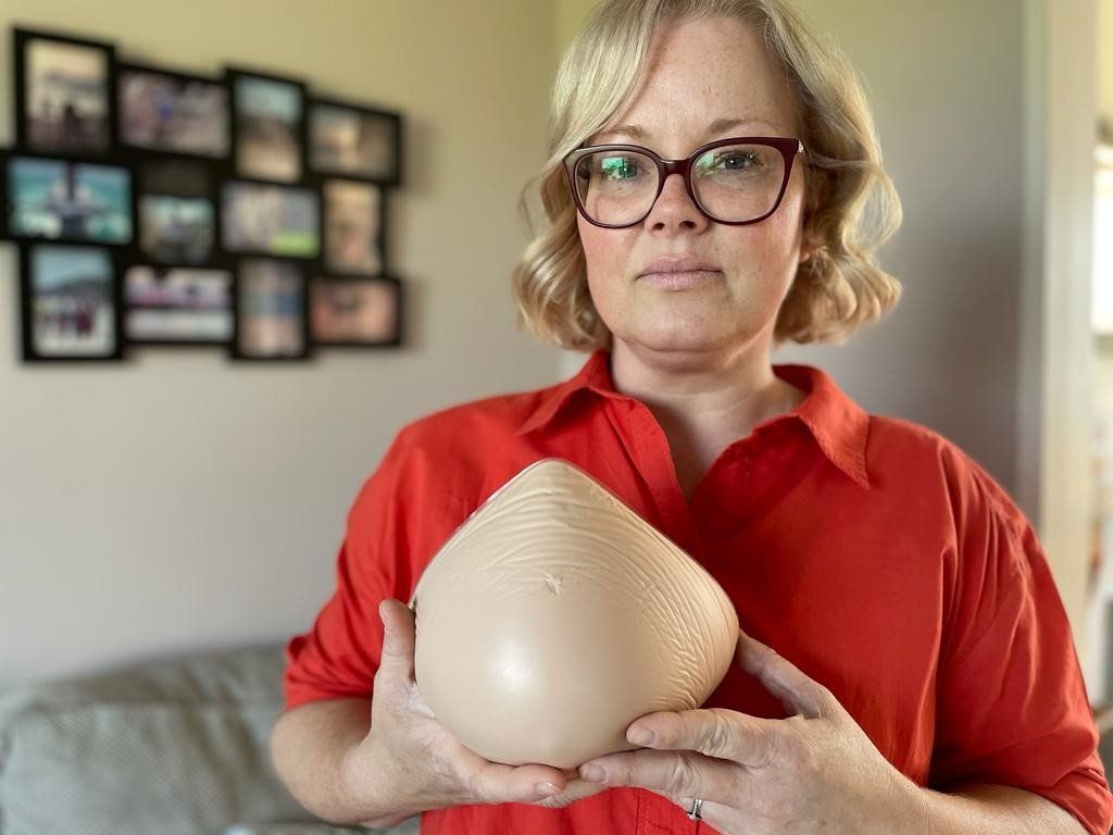 Jo has a blonde wavy bob and wears dark framed glasses. She is wearing a red shirt-dress and is holding one of her old prosthetic breasts in her hand.