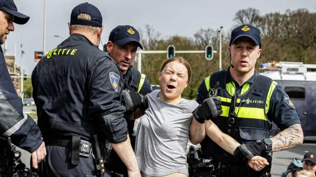 The image shows several police officers in dark uniforms with high-visibility yellow stripes detaining a person in a gray shirt. The officers wear caps and tactical gear. The scene appears to be on a street with traffic lights and trees visible in the background.