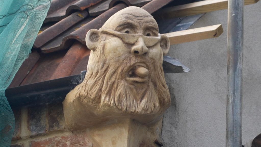 A sculpture of a man with a beard and glasses, on the edge of a roof