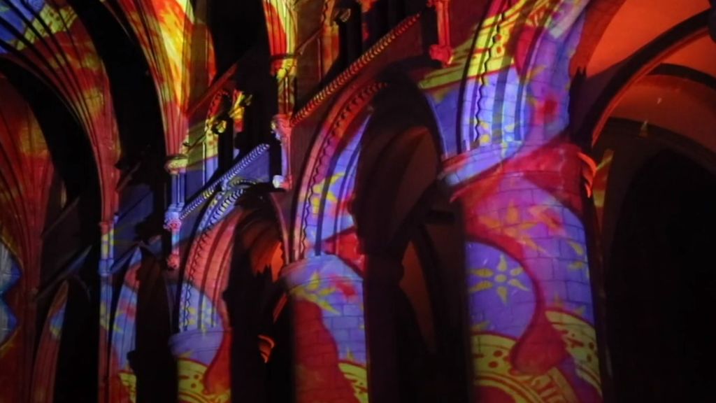 A light display of reds, blues, yellows and purples is projected onto the pillars of Gloucester Cathedral