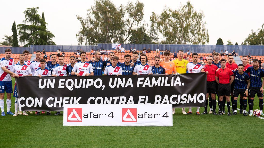 Players and officials pose for a photograph with a banner with a message against racism and in support of the goalkeeper Cheikh Kane Sarr of Rayo Majadahonda prior to the Primera RFEF Group 1 match between Rayo Majadahonda and SD Ponferradina