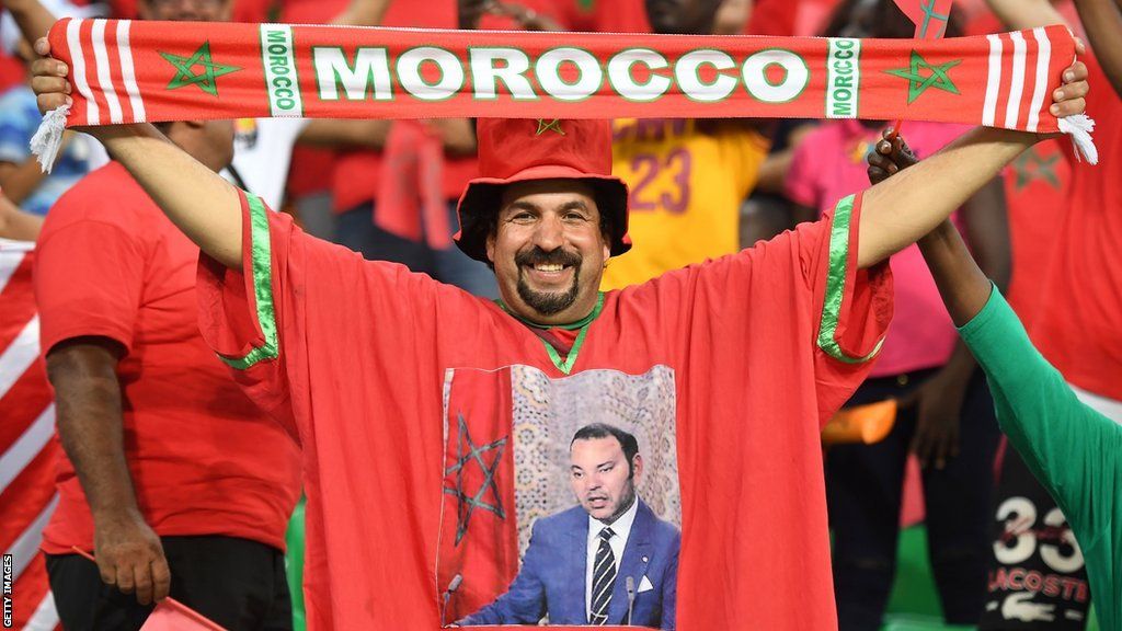 A male football fan wearing a t-shirt with a picture of King Mohammed VI on it holds up a scarf that says Morocco