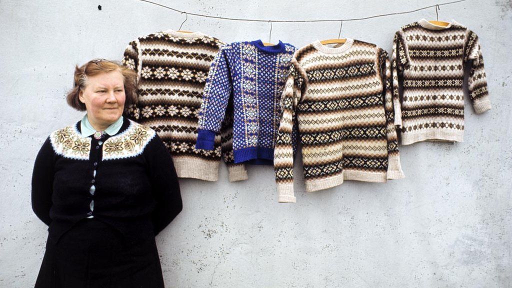 A woman poses wearing a Fair Isle style cardigan in front of Fair Isle jumpers hanging on a line in front of the wall of a cottage on one of the Shetland Islands in 1970.