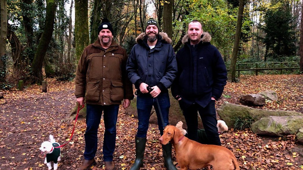 Three men looking into the camera on a dog walk with three dogs in the woods