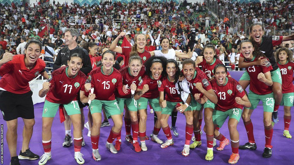 Morocco's women celebrate together at the side of a pitch with crowd behind them