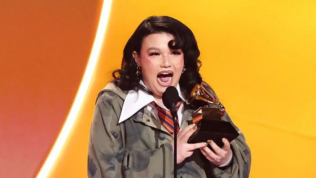Lola Young has long black hair, she is wearing a dark green jumper with a large white collar over the top, she has a orange and blue striped tie on. She is holding a GRAMMY award. 