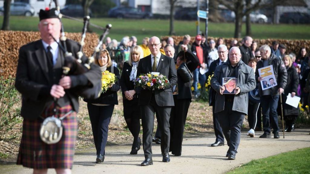 A procession of people holding flowers and signs follow a bagpiper on Glasgow Green