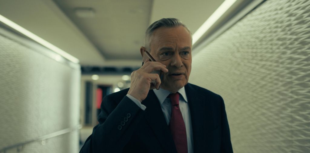 Clunes in a suit with a light blue shirt and red tie. Holding a phone to his right ear