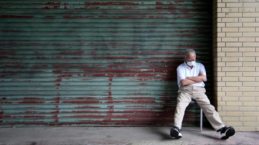 An elderly man in a white short sleeve shirt, beige trousers and black and white trainers sits with his arms folded on a chair in front of a green garage door. He appears to be taking a nap.