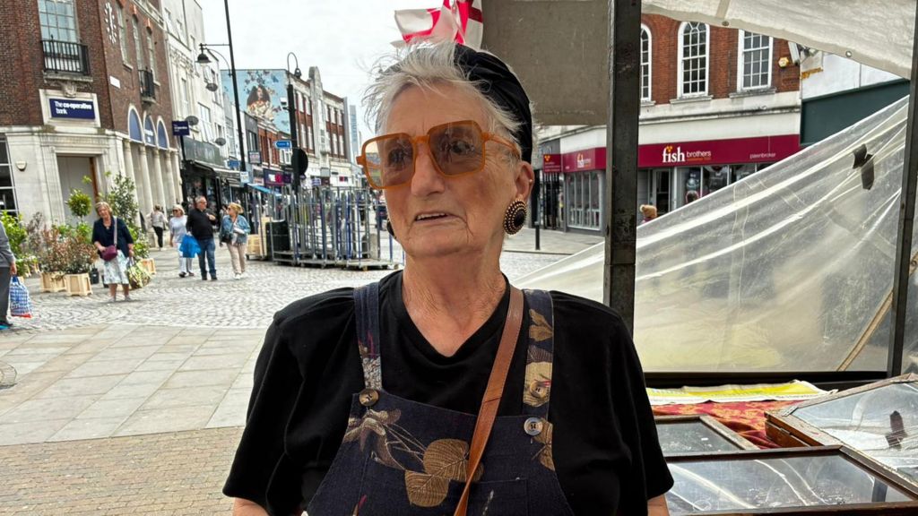 A close-up portrait of a resident named Irene standing at Romford Market. She is wearing large tinted glasses, a black top, and patterned dungarees while speaking to the camera.