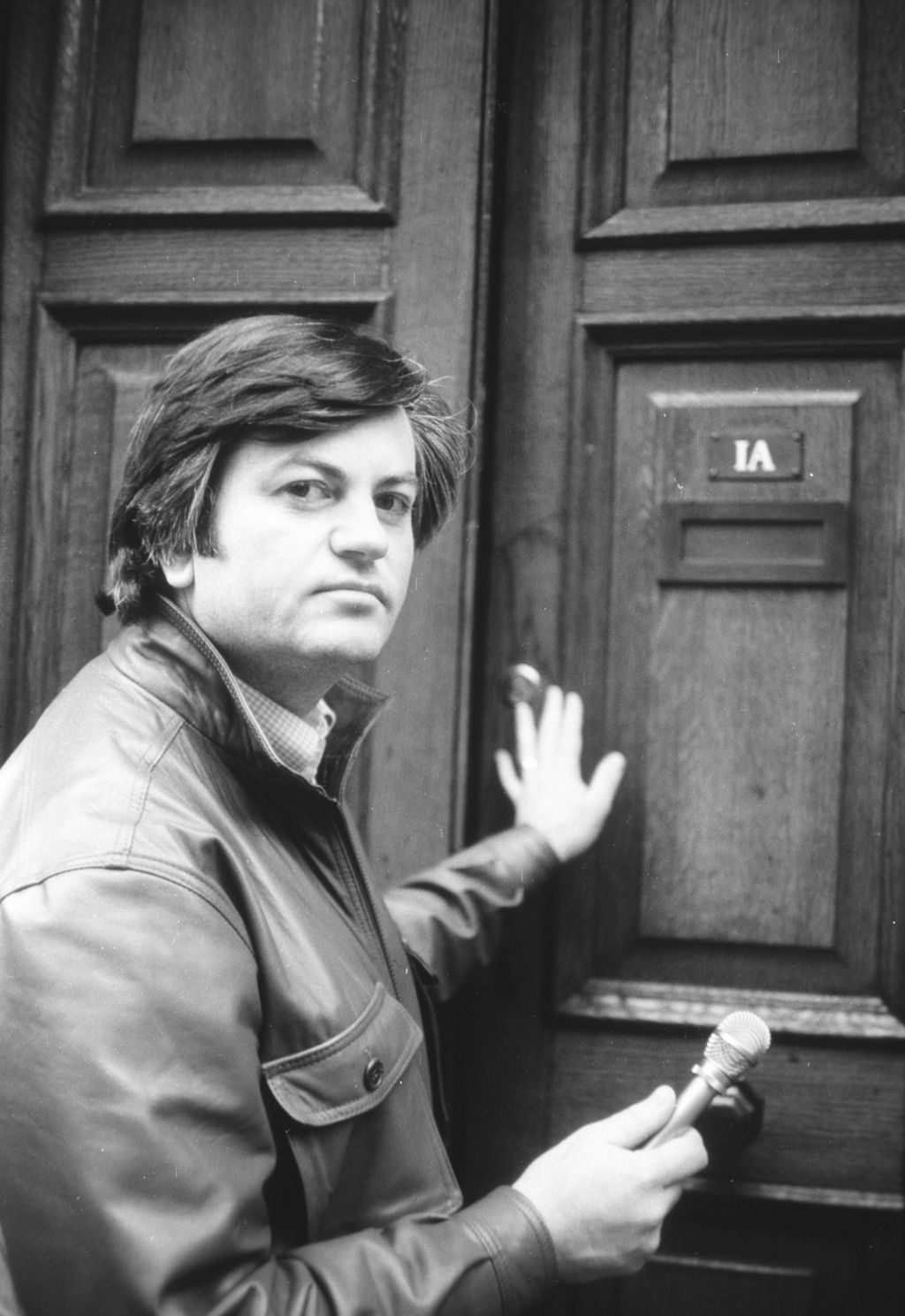 Roger Cook, pushing open a door, looking at the camera while holding a microphone