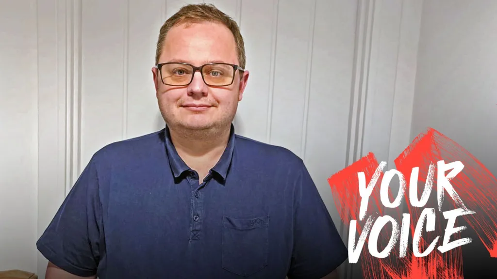 Sam Gillson, wearing glasses and a blue polo shirt, with Your Voice branding on the right hand side of the picture