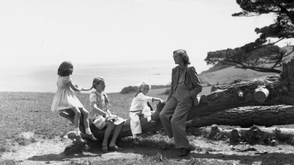 A black and white photo showing Daphne du Maurier sitting on a tree trunk with a young boy in a sailor suit and two young girls wearing dresses. Du Maurier is wearing a trouser suit. One of the girls is holding one of the other's plait. Behind them is a cliff and the sea beyond.