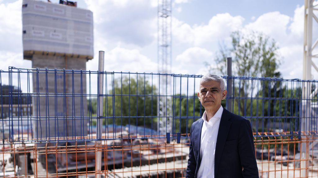 Sadiq Khan is seen a navy suit stands by metal fencing at a construction site, with cranes and a partially built structure behind him under a partly cloudy sky.