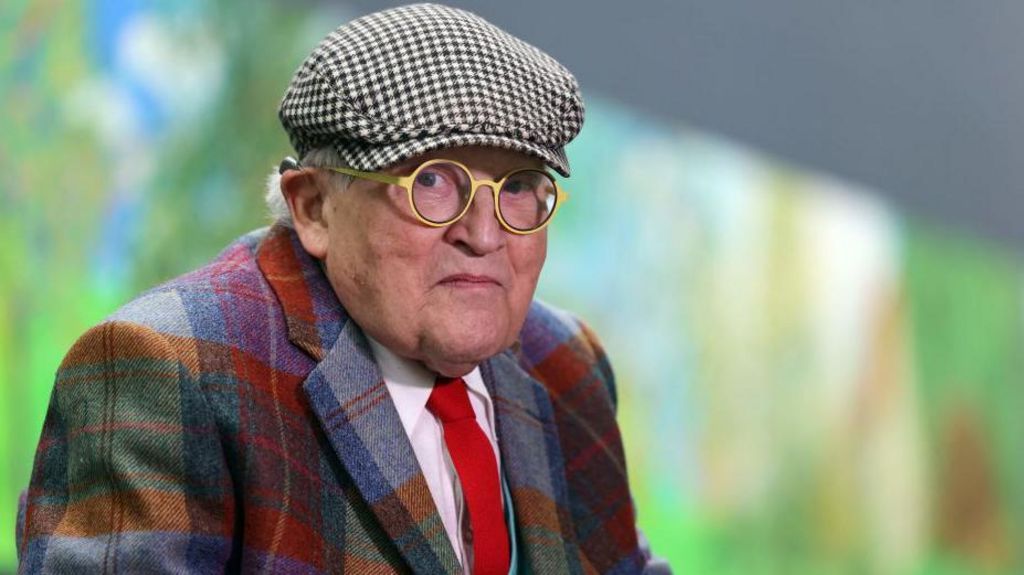 David Hockney is seen in old age. He is wearing a check suit and a check cap and yellow round glasses. He is looking into camera.