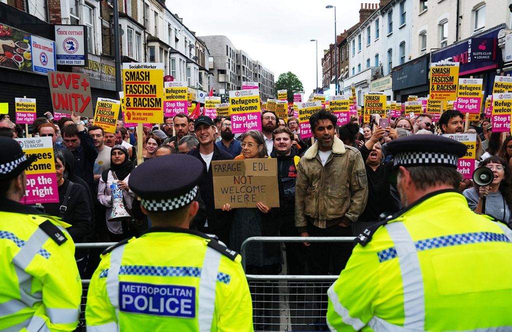 Police officers line up in front of a mass of anti-racism protesters holding placards on Hoe Street in Walthamstow on 7 August