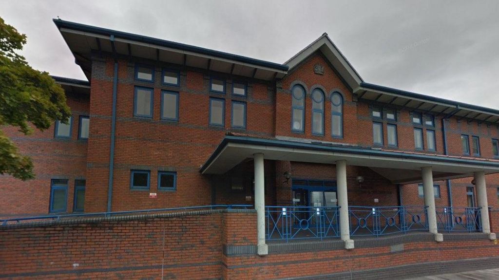 Stoke-on-Trent Court, with a ramp and blue railings visible. It is a large, two-storey red brick building with glass door behind a set of columns which hold up a tiled canopy above the entrance.