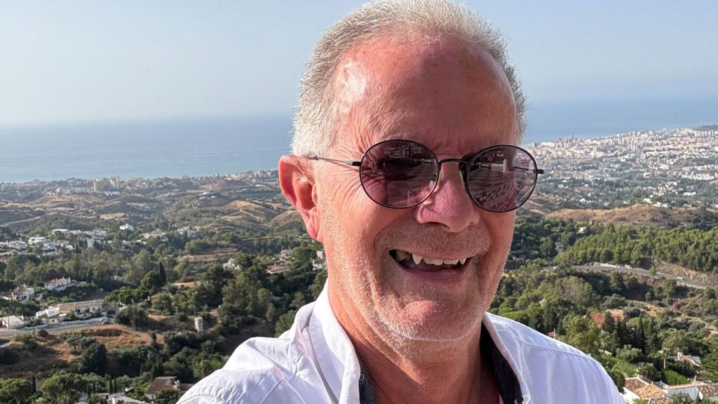 Peter Attwell smiles wearing sunglasses and a white shirt, with a background of the sea, trees and houses.